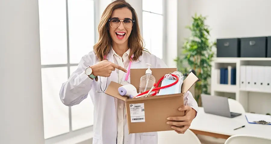 Happy female doctor holding a box filled with medical supplies and smiling while pointing.