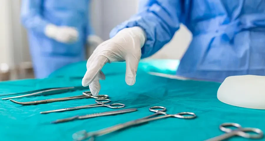 Nurse picking up a surgical instrument in an operating room, with surgeons working in the background.