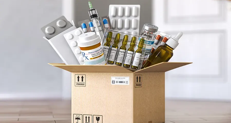 Box filled with various medical supplies including syringes, blister packs, and prescription bottles on a wooden floor.