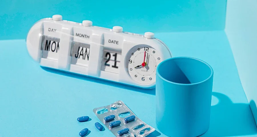 Blue cup and blister pack of pills next to a white calendar clock showing the date, representing time-sensitive medication use