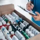 Close-up of a person inspecting a box full of assorted medical bottles and products, while taking notes on a clipboard.