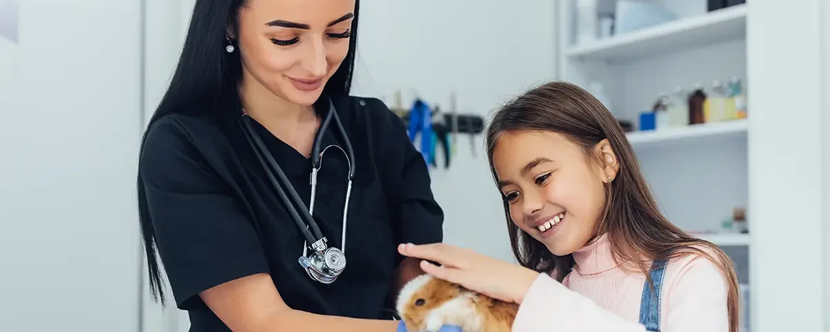 Veterinarian in black scrubs with a stethoscope examining a guinea pig while a smiling young girl watches at a veterinary clinic.