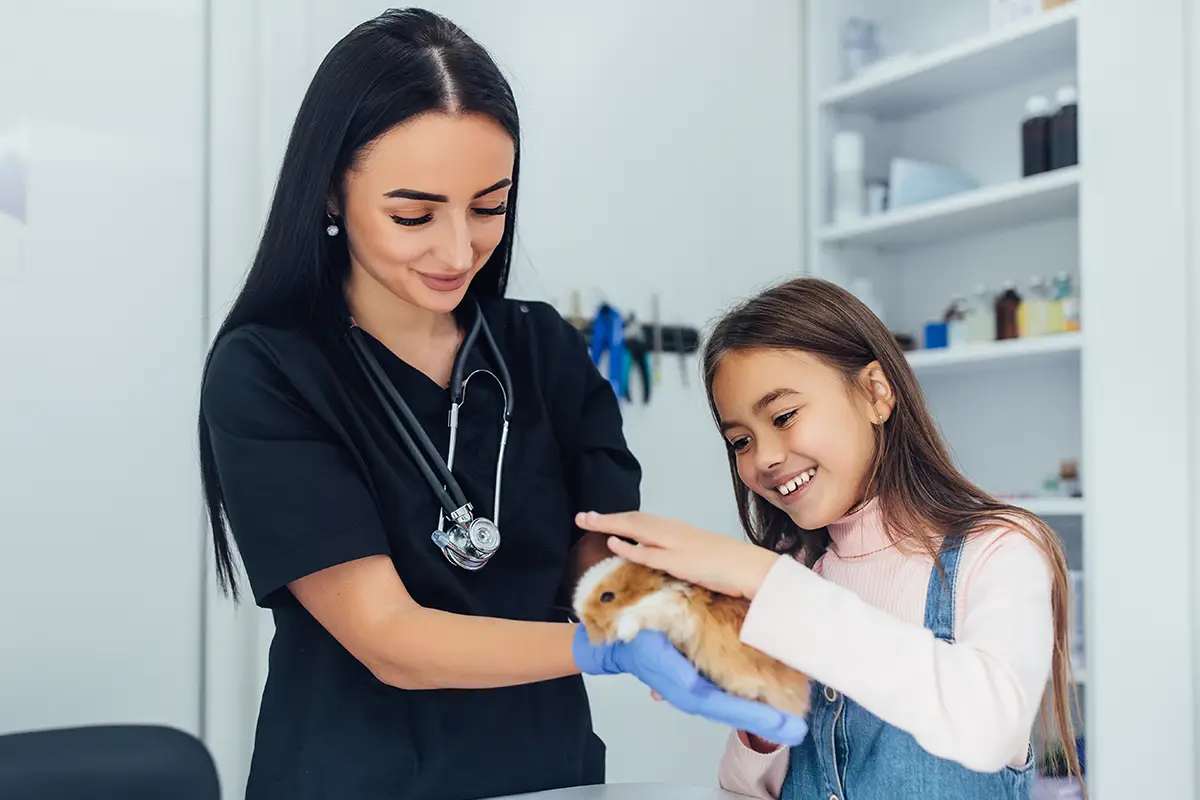 Veterinarian in black scrubs with a stethoscope examining a guinea pig while a smiling young girl watches at a veterinary clinic.